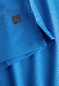 Tissu texturé bleu plié, affichant un logo triangulaire noir sur un petit patch carré près de l'ourlet.