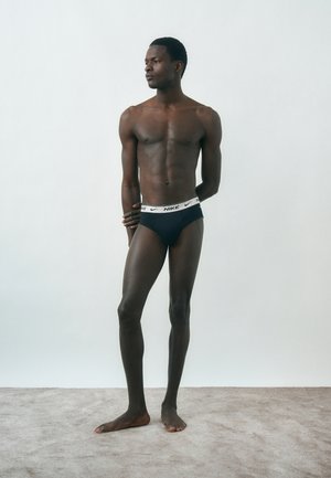 Homme athlétique à la peau foncée, debout pieds nus sur un tapis, portant un slip Nike noir avec une ceinture blanche, regardant sur le côté contre un mur blanc uni.