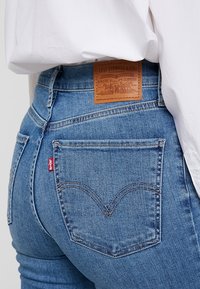 Jeans de mezclilla azul con una cintura alta, que cuentan con un parche de cuero en la parte trasera, costuras en contraste y una etiqueta roja en el bolsillo trasero.