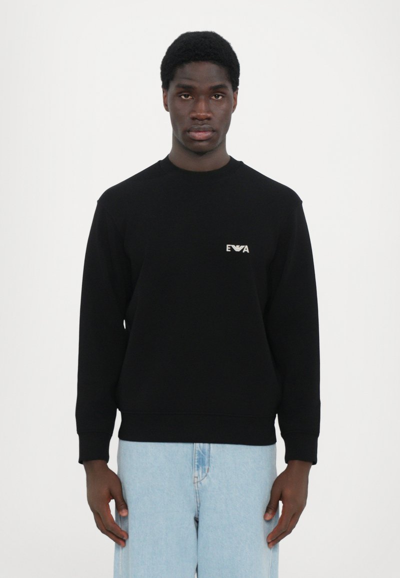 Sweatshirt noir avec col rond et poignets côtelés, arborant un petit logo blanc sur la poitrine gauche. Associé à un jean en denim bleu clair.