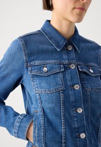 Giacca di jeans di colore blu scuro con colletto classico, chiusure con bottoni in argento e due tasche sul petto. Presenta cuciture visibili e una vestibilità strutturata.