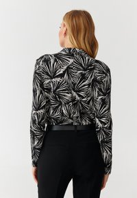 Blusa de manga larga en blanco y negro con un patrón de hojas de palma, que presenta un cuello y una parte de atrás abotonada, combinada con pantalones negros de cintura alta.