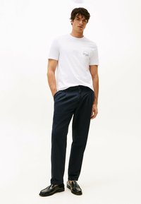Camisa de manga corta blanca con un bolsillo en el pecho, combinada con pantalones oscuros de color navy y mocasines negros, sobre un fondo blanco liso.