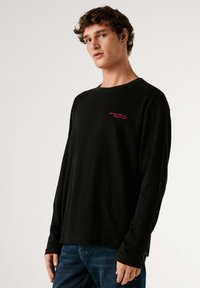 Črna majica z dolgimi rokavi z rdečim vezenim napisom na prsih. Mehka tkanina z sproščenim krojem in okroglim izrezom. Oblečena z modrimi kavbojkami.