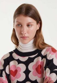 Mujer joven con cabello castaño claro que lleva un suéter negro con grandes patrones de flores rosas y rojas y un cuello de tortuga blanco debajo.