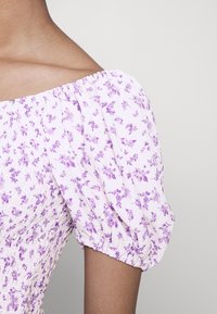 Primo piano di una spalla e manica di un vestito con motivo floreale, caratterizzato da un tessuto rosa chiaro con fiori viola e maniche a sbuffo arricciate.