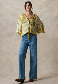 Bluză fluidă, galbenă deschis, cu un model floral, asociată cu blugi largi, blue, și cizme de gleznă cu toc maro. Textura naturală a materialului este vizibilă.