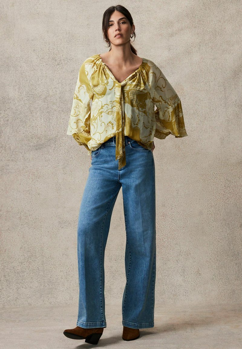 Bluză fluidă, galbenă deschis, cu un model floral, asociată cu blugi largi, blue, și cizme de gleznă cu toc maro. Textura naturală a materialului este vizibilă.