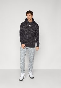 Mand iført sort mønstret hættejakke og lys grå zigzag-mønstret joggingbukser med hvide sneakers, stående foran en ensfarvet baggrund.
