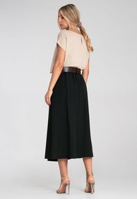 Beige Cropped-Top mit kurzen Ärmeln, kombiniert mit einem langen schwarzen Rock, der einen breiten braunen Gürtel trägt. Transparente Sandalen mit Absatz runden den Look ab.