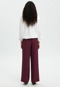 Femme aux longs cheveux bouclés vue de dos, portant une blouse blanche, un pantalon large bordeaux et des talons noirs pointus.