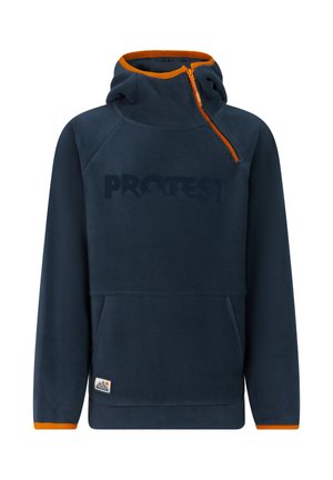 Jongen met een marineblauwe capuchonjas, oranje muts en oranje broek, staand met één hand in zijn zak voor een lichte achtergrond.