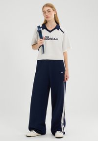 Kobieta w białej i granatowej koszulce polo oraz granatowych spodniach z szerokimi nogawkami w białe paski, trzymająca pasek granatowego plecaka na jednym ramieniu.