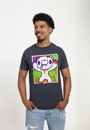 Mężczyzna o krótkich kręconych włosach, ubrany w ciemnoszary t-shirt z kolorową grafiką kreskówkowego misia oraz jasnoniebieskie, podarte dżinsy, stoi z rękami w kieszeniach.