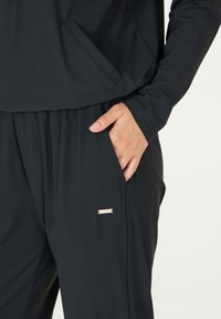 Schwarzes Sportoutfit mit lässiger Passform, ausgestattet mit einer Fronttasche und mit goldfarbenen Akzenten an der Hose. Glatte, weiche Stofftextur.