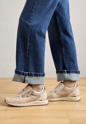 Jalat, joissa beige-tossut sivuvetoketjuilla ja beige-sukat, yhdistettynä käännettyihin sinisiin farkkuihin, seisovat puulattialla.