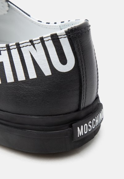 Φτέρνα μαύρου δερμάτινου αθλητικού παπουτσιού με μεγάλο λευκό μερικό κείμενο «MOSCHINO» και μικρότερο «MOSCHINO» στην ετικέτα της μαύρης ελαστικής σόλας.