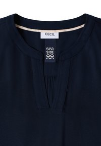 Blouse bleu marine avec un col rond, un détail plissé à l'avant et un col intérieur contrastant de couleur claire. Étiquettes "CECIL" et "sea SIDE" visibles.