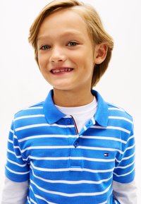 Αγόρι που χαμογελάει με ανοιχτό καστανά μαλλιά, φορώντας μπλε και λευκή ριγέ πόλο μπλούζα πάνω από λευκό μακρυμάνικο πουκάμισο, μπροστά σε απλό φόντο.
