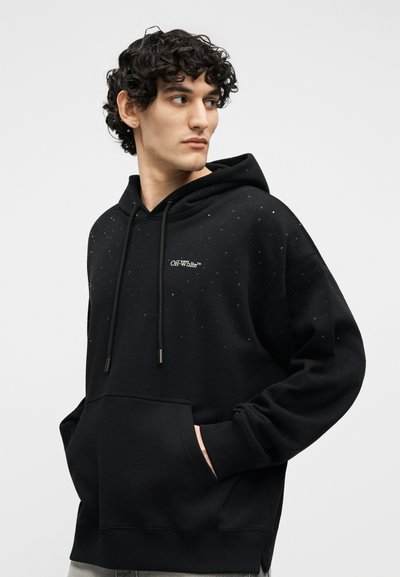 Jauns vīrietis ar cirtainiem matiem, valkājot melnu Off-White kapuci ar izkaisītiem baltiem punktiem, rokas priekšējā kabatā, skatās sāniski.
