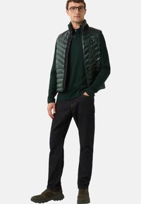 Dunkelgrüne Steppweste über einem schwarzen Langarm-Poloshirt, kombiniert mit dunklen Jeans und braunen Sneakers. Ausgestattet mit strukturierter Stoffoberfläche und feinen Nähten.