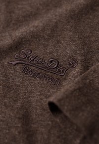 Brun bomuldsstof med en blød tekstur, prydet med broderet "SuperDry" logo i en mørkere brun nuance. Nærbillede af materialet.
