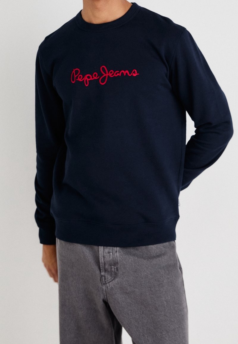 Homme portant un sweat-shirt bleu marine avec le logo rouge "Pepe Jeans" et un jean gris, debout avec une main derrière le dos.