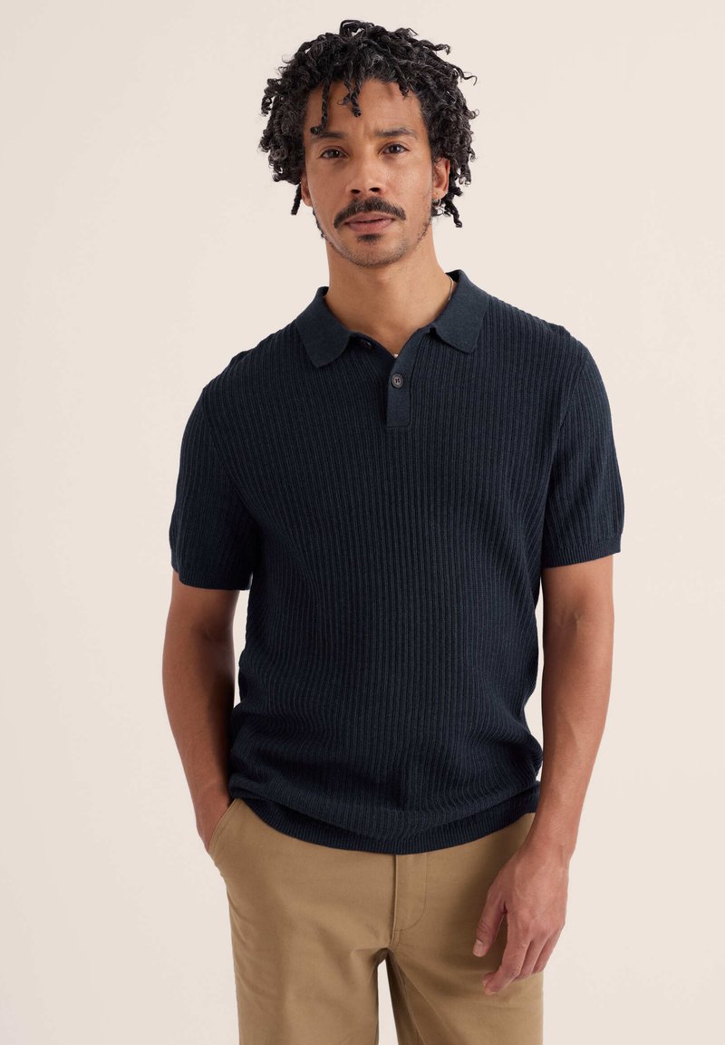Homme aux cheveux bouclés et moustache portant un polo à manches courtes côtelé foncé et un pantalon marron clair, debout avec une main dans la poche.