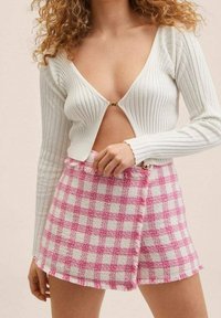 Vrouw draagt een wit geribd langermig shirt met een sluiting aan de voorkant en een hoog getailleerde roze-wit geruite rok.