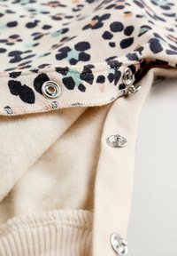 Tissu beige clair avec des taches de léopard noires, vert menthe et pêche, comportant des fermetures à pression en métal sur le bord et un intérieur doux et doublé.