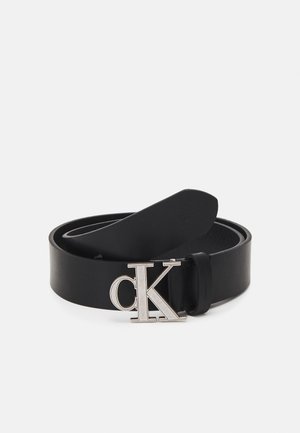 Ceinture en cuir noir enroulée avec boucle métallique argentée entrelacée "cK" sur un fond gris clair uni.