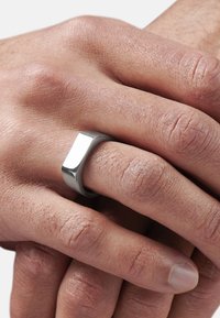 Metallring med rektangulært topp, glatt tekstur og matt finish. Bæres på en hånd som viser lett teksturert hud.