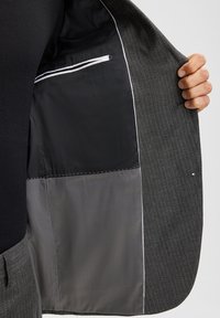 Hand, die eine offene graue Nadelstreifenanzugjacke hält, die mit einem schwarz-grauen Innenfutter und einer weiß eingefassten Innentasche ausgestattet ist.