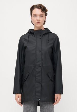 Femme portant un imperméable noir à capuche avec des boutons à l'avant et des poches à rabat inclinées, se tenant contre un fond blanc uni.
