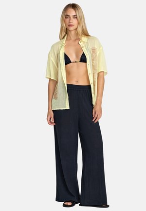 Camicia a maniche corte giallo chiaro con tessuto a maglia aperta e dettagli decorativi, abbinata a pantaloni neri a gamba larga e un bikini nero.