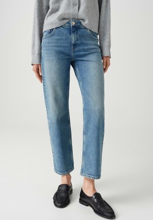 Frau trägt hellblaue gerade geschnittene Jeans, grauen Pullover und schwarze Ledermokassins und steht vor einem schlichten hellen Hintergrund.