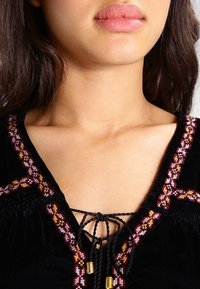 Blouse en velours noir avec une broderie complexe rouge et jaune le long de l'encolure. Présente un devant à lacets avec des pampilles et des accentuations dorées.