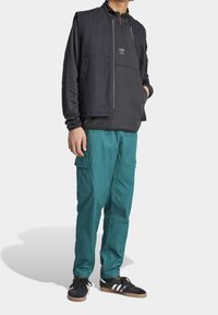 Svart zip-up väst över en mörkgrå tröja, parat med teal cargobyxor. Svarta sneakers med vita ränder fullbordar looken.