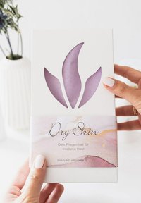 Weißer Verpackungsbehälter mit einem lila floralen Ausschnitt. Auf dem Etikett an der Vorderseite steht der Text "Trockene Haut" und der Hintergrund ist in einer strukturierten Aquarell-Optik in Pink und Gold gestaltet.