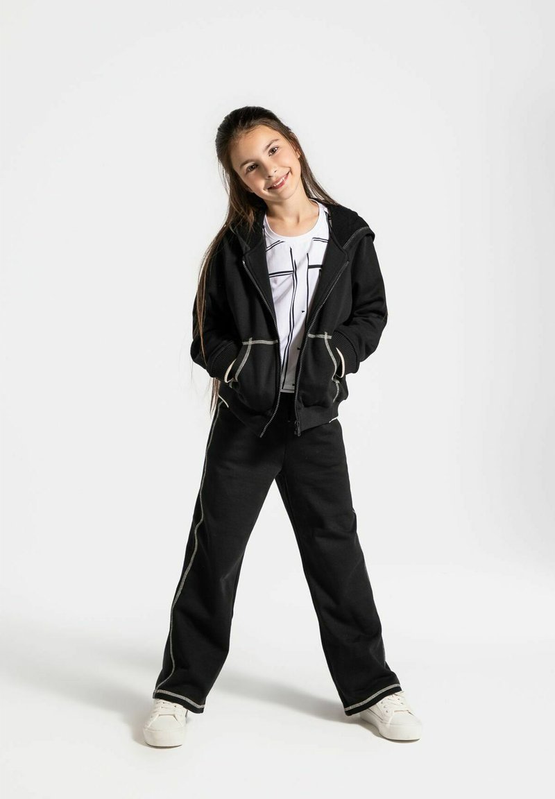 Czarna bluza z zamkiem błyskawicznym z jasnoszarymi akcentami, zestawiona z czarnymi spodniami z podobnymi detalami. Stylizowana z białą koszulą i białymi trampkami.