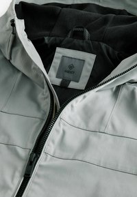 Jachetă crem cu fermoar, căptușită cu un fleece moale negru, având un patch cu logo și detalii cusute structurale pe exterior.
