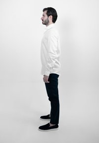 Witte blouse met lange mouwen, zwarte broek, zwarte instappers. Onderwerp staat zijwaarts tegen een plain witte achtergrond. Minimalistisch ontwerp.