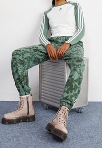 Person som sitter på en silverfärgad metallåda med hjul, klädd i gröna tie-dye sweatpants, en vit och grön Adidas långärmad tröja och beige snörstövlar.