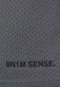 Tissu en maille gris avec de petites perforations, mettant en avant le texte "9N1M SENSE" brodé en noir. Texture lisse avec un design respirant.