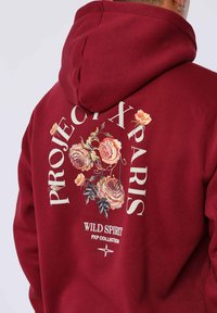 Sudadera roja con diseños florales en la parte posterior, con el texto "PROJECT X PARIS" y "WILD SPIRIT". El material es suave y tiene un corte relajado.