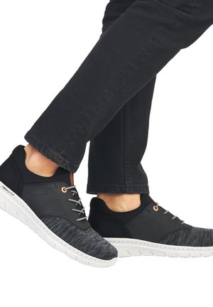 Des chaussures de sport noires avec des accents en textile gris, présentant un dessus lisse, une semelle blanche et des lacets gris, positionnées dans un mouvement de marche.