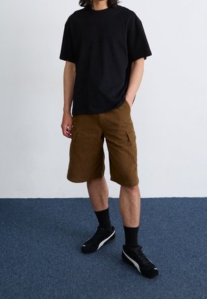 Osoba ubrana w czarną koszulkę z krótkim rękawem, brązowe szorty typu cargo, czarne wysokie skarpetki i czarne trampki, stojąca na niebieskim dywanie.