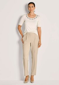 Vrouw staat met haar hand in de zak, draagt een crèmekleurig bewerkt topje met een ronde hals, beige op maat gemaakte broek en beige hoge hakken tegen een witte achtergrond.