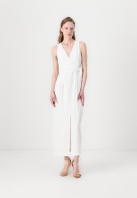 Rochie albă cu decolteu adânc în V, fără mâneci, cu fente în față și material fluid. Asortată cu sandale bej cu toc înalt.