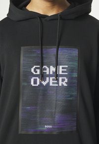 Sweat à capuche noir présentant un graphisme rectangulaire avec l'inscription "GAME OVER" en style pixel art, un fond texturé violet et vert, et le nom de la marque "BOSS".